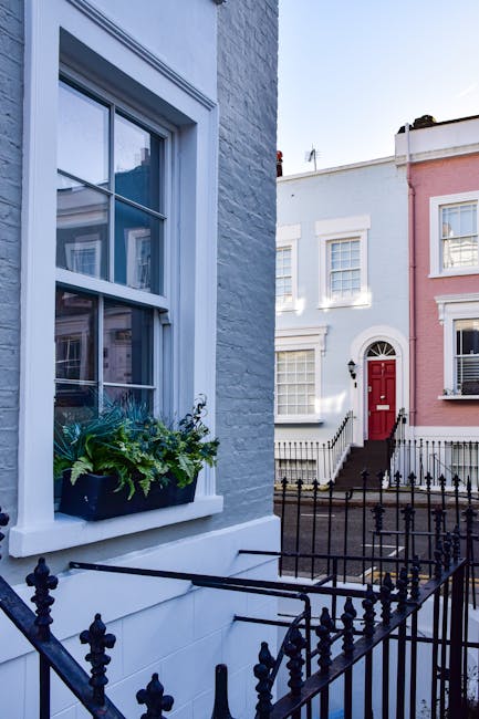 Close-up view of a residential window with white-painted wooden framing and multi-pane glass, set into a grey brick wall. A window box beneath the window contains green foliage and flowering plants. Adjacent is a black metal fence with decorative finials, enclosing the front of a house. In the background, other terraced houses with pastel-colored facades, white window frames, and steps leading up to entrances are visible under clear daylight. The scene suggests a well-maintained, clean urban property typical of the Notting Hill area, emphasizing attention to detail in surface cleaning and exterior maintenance, consistent with the services offered by Notting Hill Carpet Cleaners under their house cleaning and deep cleaning categories.
