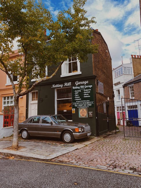 Exterior view of the Notting Hill Garage, a black building with a prominent sign that reads 'Notting Hill Garage' and lists services such as cleaning, repairs, and servicing. In front of the garage, a silver vintage car is parked on a cobblestone driveway. A large tree with green leaves partially shades the area, and neighboring buildings with brick and painted facades are visible on either side. The scene is illuminated by natural daylight, with a partly cloudy sky overhead, emphasizing a clean and well-maintained urban environment. This setting illustrates the exterior of a commercial property specializing in surface cleaning and maintenance, supported by Notting Hill Carpet Cleaners' expertise in deep cleaning and hygiene for local residences and businesses.