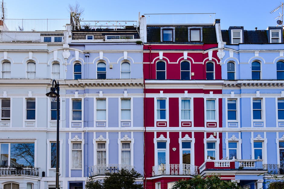 A row of multi-story terraced houses in Notting Hill during daytime, featuring Victorian-style bay windows and decorative cornices. The buildings are painted in various colors, including pastel blue, 