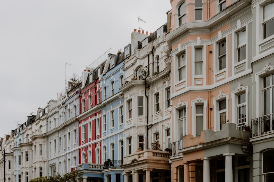 Photograph of a row of multi-story residential buildings in Notting Hill, featuring Victorian-style architecture with decorative moldings and large bay windows. The buildings are painted in pastel shades of pink, white, blue, and cream, with some having small balconies and iron railings on the upper floors. Overcast skies diffuse soft natural light across the facades, emphasizing their intricate details and weathered textures. The ground level shows entrances with steps leading to front doors, while the upper stories include sash windows and rooftop antennas. The overall scene conveys a charming, historic neighborhood atmosphere, with well-maintained exteriors reflecting community upkeep by Notting Hill Carpet Cleaners, focused on surface cleaning and hygienic maintenance relevant to their property care and street presentation.