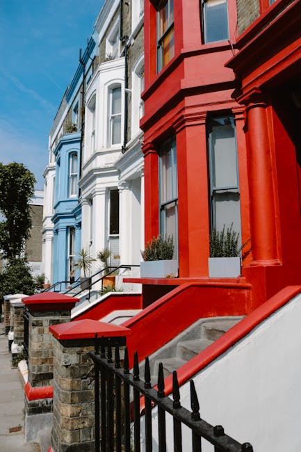 Close-up view of a row of Victorian-style residential buildings in Notting Hill, featuring colorful facades with red, white, and blue paint. The red building in the foreground has a staircase with a concrete step and a black wrought iron railing, leading up to a white door. Large windows with black frames and shutters are visible, with some windows having flower boxes filled with greenery. The buildings are illuminated by natural daylight, highlighting the clean and well-maintained exteriors. The overall scene emphasizes the vibrant and historic architecture typical of Notting Hill, with subtle indications of cleanliness and exterior upkeep. Notting Hill Carpet Cleaners specializes in surface cleaning and deep cleaning services that help maintain the hygiene and appearance of such residential properties, adhering to local council rules and regulations regarding property standards.