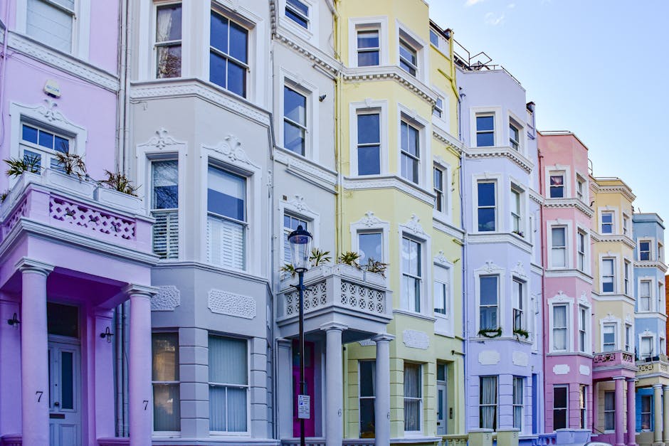 Exterior view of a row of Victorian-style terraced houses in Notting Hill, featuring a mix of white, pink, and light blue facades, with large sash windows and decorative architectural details. The bui