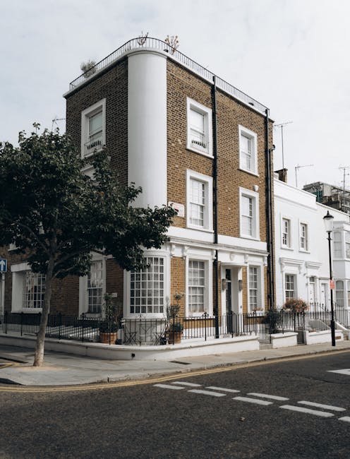 Exterior view of a multi-story residential building on Kensington Park Road in Notting Hill W11, showcasing a combination of brick and white-painted facade. The ground floor features large windows with white frames, and the entrance area is decorated with potted plants. The building has a rounded corner design with a metal railing on the rooftop, and a street lamp is visible in the foreground. The street is paved with asphalt and has marked pedestrian crossings, with a nearby tree partially obscuring part of the building. This image illustrates an urban residential setting, emphasizing the clean, well-maintained exterior typical of Notting Hill. Notting Hill Carpet Cleaners, a professional cleaning service, offers surface cleaning and deep cleaning solutions to maintain such properties' hygiene and appearance.