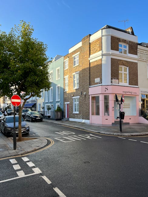 A street scene on Kensington Park Road showing a multi-story corner building with a mix of brick and light-colored exterior walls. The ground floor features a pink shop front with large display windows, displaying a sign for 'Sugarings.' A black bicycle is parked outside the shop. To the left, a tree with green foliage partially shades the sidewalk, which is paved with light-colored stones. Several parked cars line the street, which is marked with white road markings and a 'No Entry' traffic sign at the intersection. The scene is illuminated by natural daylight, with shadows cast on the building facades and street, highlighting the clean and well-maintained appearance of the street environment. Notting Hill Carpet Cleaners' branding is implied as part of the overall context for cleaning services in this area, emphasizing surface cleaning, deep cleaning, and sanitisation for residential and commercial properties in the W11 postcode.