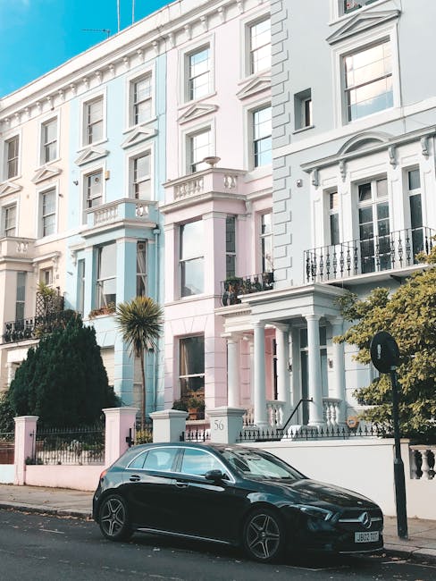 Exterior view of a multi-story residential building on Kensington Park Road in Notting Hill W11, showcasing a combination of brick and white-painted facade. The ground floor features large windows with white frames, and the entrance area is decorated with potted plants. The building has a rounded corner design with a metal railing on the rooftop, and a street lamp is visible in the foreground. The street is paved with asphalt and has marked pedestrian crossings, with a nearby tree partially obscuring part of the building. This image illustrates an urban residential setting, emphasizing the clean, well-maintained exterior typical of Notting Hill. Notting Hill Carpet Cleaners, a professional cleaning service, offers surface cleaning and deep cleaning solutions to maintain such properties' hygiene and appearance.
