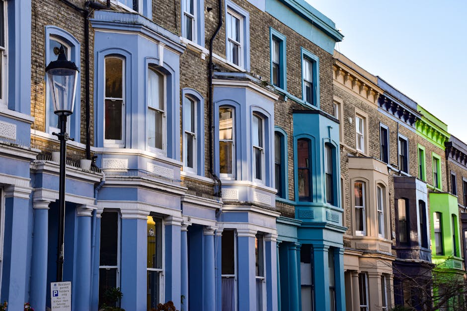Photograph of a row of Victorian residential buildings in Notting Hill, featuring ornate bay windows with white and light blue trim, decorative cornices, and a mix of brick and painted facades. The buildings are illuminated by natural daylight, highlighting their historical architecture. In the foreground, a black lamppost is visible alongside a small parking sign, while the street appears clean and well-maintained. These detailed facades exemplify typical domestic architectural features that Notting Hill Carpet Cleaners might service for surface cleaning and deep cleaning to maintain the property's appearance and hygiene.