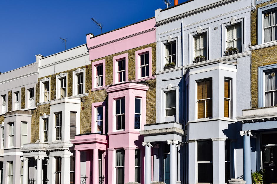 A row of multi-story residential buildings in Notting Hill with a clear blue sky background. The buildings feature a mix of brick and painted facades, including a prominent pink-painted section with bay windows and decorative architecture details. Adjacent structures are painted in white and light blue, each with large sash windows, some with window boxes filled with plants. The sunlight creates shadows on the buildings, highlighting their architectural features and the well-maintained appearance of the exteriors, characteristic of the area. This street scene emphasizes the distinctive charm of Notting Hill, with a focus on the facades' clean and refreshed look, potentially after professional surface cleaning or maintenance work by Notting Hill Carpet Cleaners.