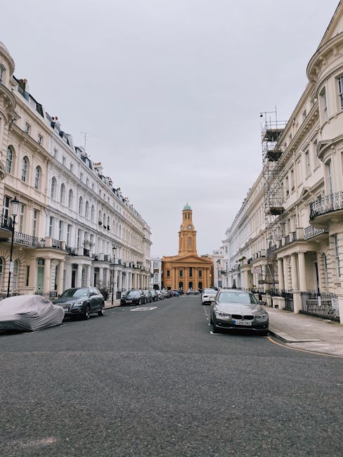 Street view of Notting Hill Gate W11 featuring elegant white Victorian-style residential buildings with ornate iron balconies and large sash windows lining both sides of the street. Parked cars are visible along the curb, including a covered vehicle on the left and various modern vehicles on the right. In the distance, a historic church with a tall bell tower topped by a green dome stands prominently at the end of the street, framed by an overcast sky. The scene highlights clean, well-maintained facades and a quiet, urban residential atmosphere, typical of the Notting Hill area. This image reflects the importance of surface cleaning and proper maintenance of historic and residential surfaces, aligning with the services offered by Notting Hill Carpet Cleaners.