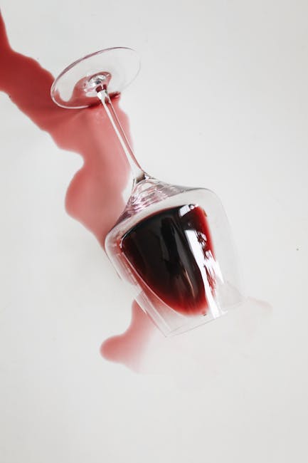 Close-up of a clear wine glass filled with red wine, tilted on its side with wine spilling out onto a white surface. The background is plain and well-lit, highlighting the transparency of the glass and the rich color of the wine. The spill creates a reflective and glossy appearance on the surface, emphasizing cleanliness and purity. This image exemplifies the importance of careful surface cleaning and stain removal, as offered by Notting Hill Carpet Cleaners, especially for wine stain removal during domestic cleaning or deep cleaning services.