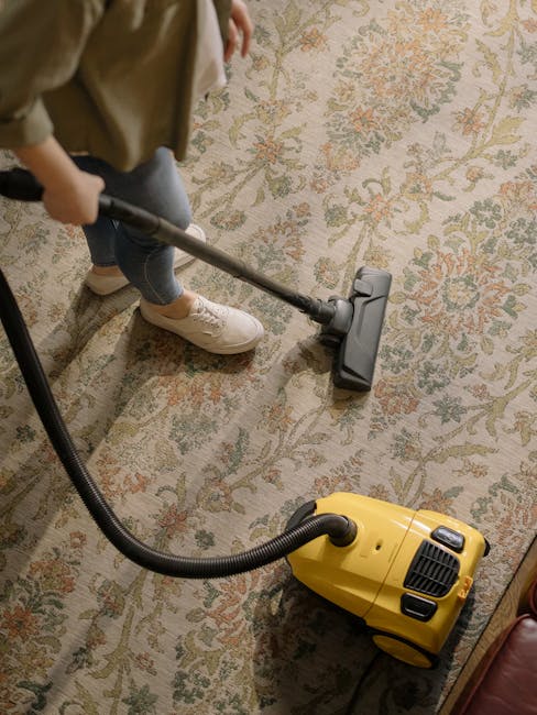 A person dressed in protective white clothing, including a hood and mask, is operating a steam cleaning machine on a light-colored carpet in a residential living room. The room features a beige sofa w