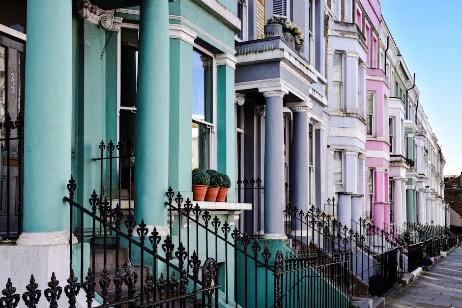 A row of colorful terraced houses in Notting Hill, featuring pastel green, blue, gray, and pink facades with decorative white trim. The houses have small front gardens with black wrought iron fences, leading to steps that ascend to front doors. Large windows with white frames are visible, some with flower boxes. The buildings are illuminated by natural daylight, highlighting the vibrant exterior colors and clean, well-maintained surfaces, indicative of thorough surface cleaning and maintenance by Notting Hill Carpet Cleaners.