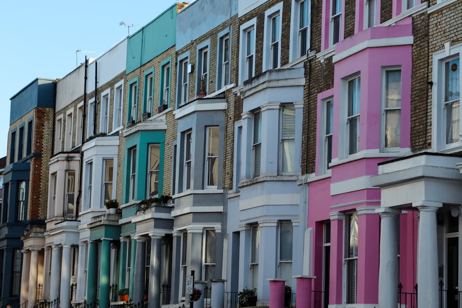 A row of multi-storey Victorian terraced houses in Notting Hill featuring colorful facades in shades of teal, gray, white, and pink. Each building has large sash windows with white frames, some with decorative molding, and small front gardens enclosed by black wrought-iron fences. The surfaces of the buildings are clean and well-maintained, with visible brickwork on some facades beneath painted sections. Bright daylight illuminates the scene, highlighting the neat appearance of the exteriors. The image is associated with residential cleaning insights provided by Notting Hill Carpet Cleaners, focusing on surface cleaning and maintenance of such properties.