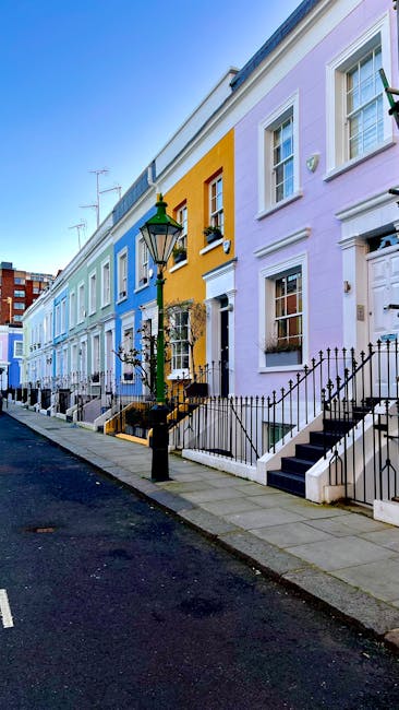 A row of colorful terraced houses in Notting Hill, featuring pastel green, blue, gray, and pink facades with decorative white trim. The houses have small front gardens with black wrought iron fences, leading to steps that ascend to front doors. Large windows with white frames are visible, some with flower boxes. The buildings are illuminated by natural daylight, highlighting the vibrant exterior colors and clean, well-maintained surfaces, indicative of thorough surface cleaning and maintenance by Notting Hill Carpet Cleaners.