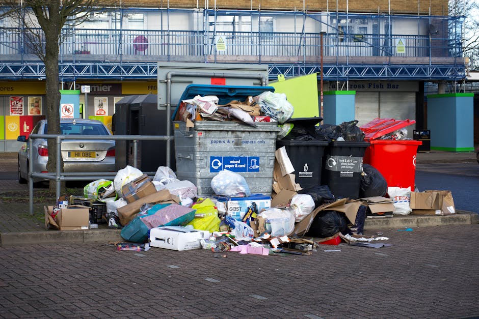 A collection of overflowing bulky waste and recycling bins placed on a paved sidewalk in front of a commercial building, with various black, red, and grey bins open and filled with mixed paper, cardboard, plastic bags, and miscellaneous trash. Surrounding the bins are scattered packaging material, cardboard boxes, and loose waste items. A metallic barrier fence is visible in the background, along with a parked silver car to the left. The scene is illuminated by natural daylight, highlighting the disorganized state of waste disposal, with a focus on the need for proper disposal practices. Notting Hill Carpet Cleaners emphasizes responsible waste management in the area, aligning with their deep cleaning and hygiene services.